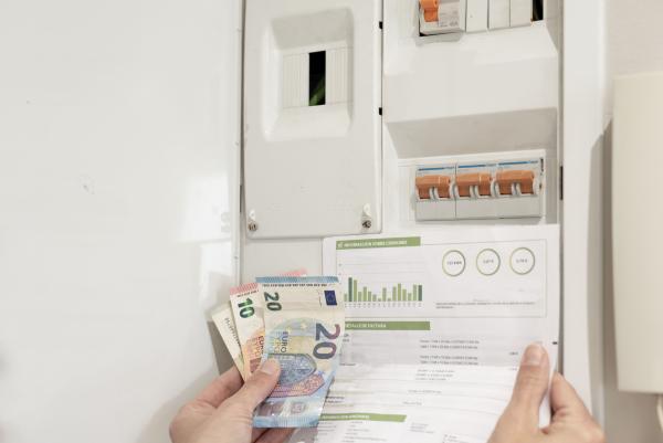 eine Hand, die die Stromrechnung und die Euro-Banknoten vor dem Netzschalter hält