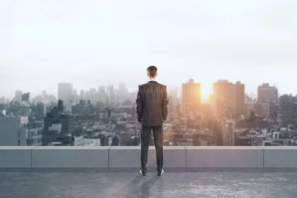 Rückansicht eines jungen Geschäftsmannes auf dem Dach mit Blick in die Ferne auf Stadthintergrund mit
