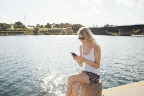 young woman sitting at riverside texting on smartp 2022 03 08 00 06 06 utc